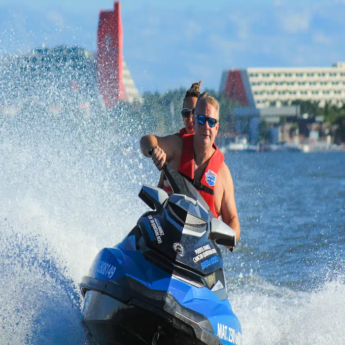 Happy couple riding a fast jet ski and making a splash on the Cancun Lagoon with the Hotel Zone in the background.